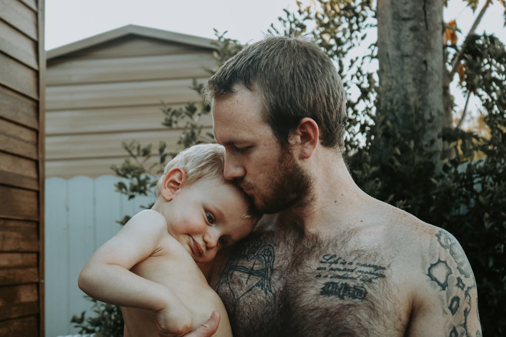 A father lovingly kisses his son on the forehead in a garden. Tattoos visible.