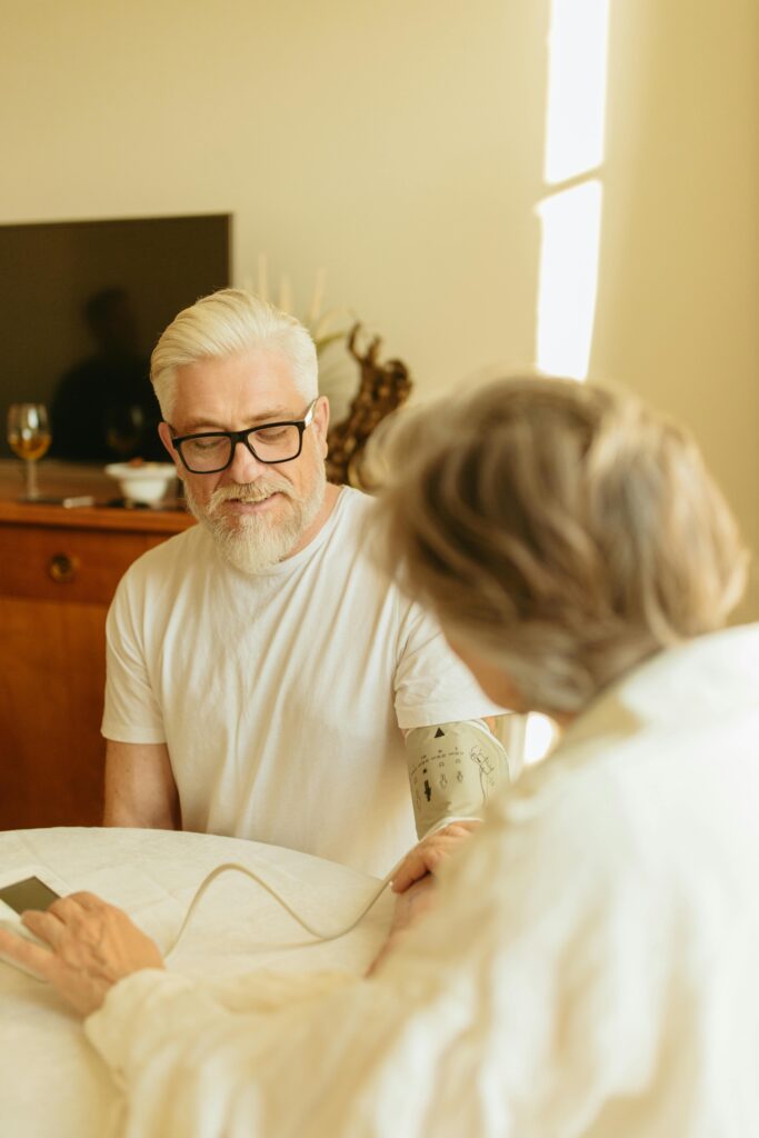Senior man with gray hair and glasses checking blood pressure using a digital monitor at home.