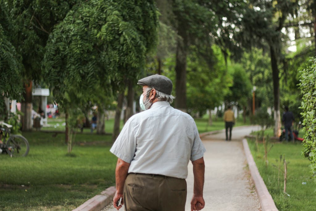 Elderly man wearing mask strolling in a lush green park, depicting pandemic lifestyle.